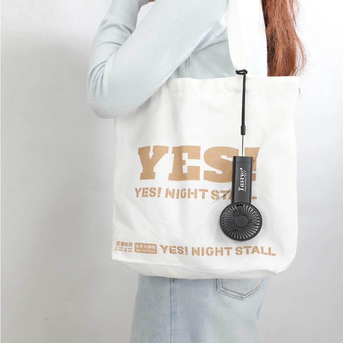 Easy Breeze hand fan clipped by its wrist strap to a canvas tote bag, lifestyle carry shot