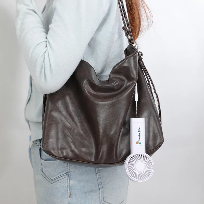 Easy Breeze hand fan clipped to a leather shoulder bag via its wrist strap, lifestyle carry shot